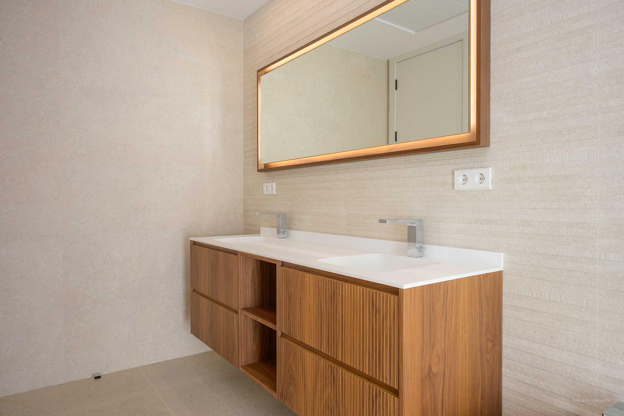 Modernes schwebendes Holzwaschtisch mit zwei integrierten Waschbecken und Chromarmaturen. Beleuchteter Spiegel mit Holzrahmen und strukturierte Wandfliesen.