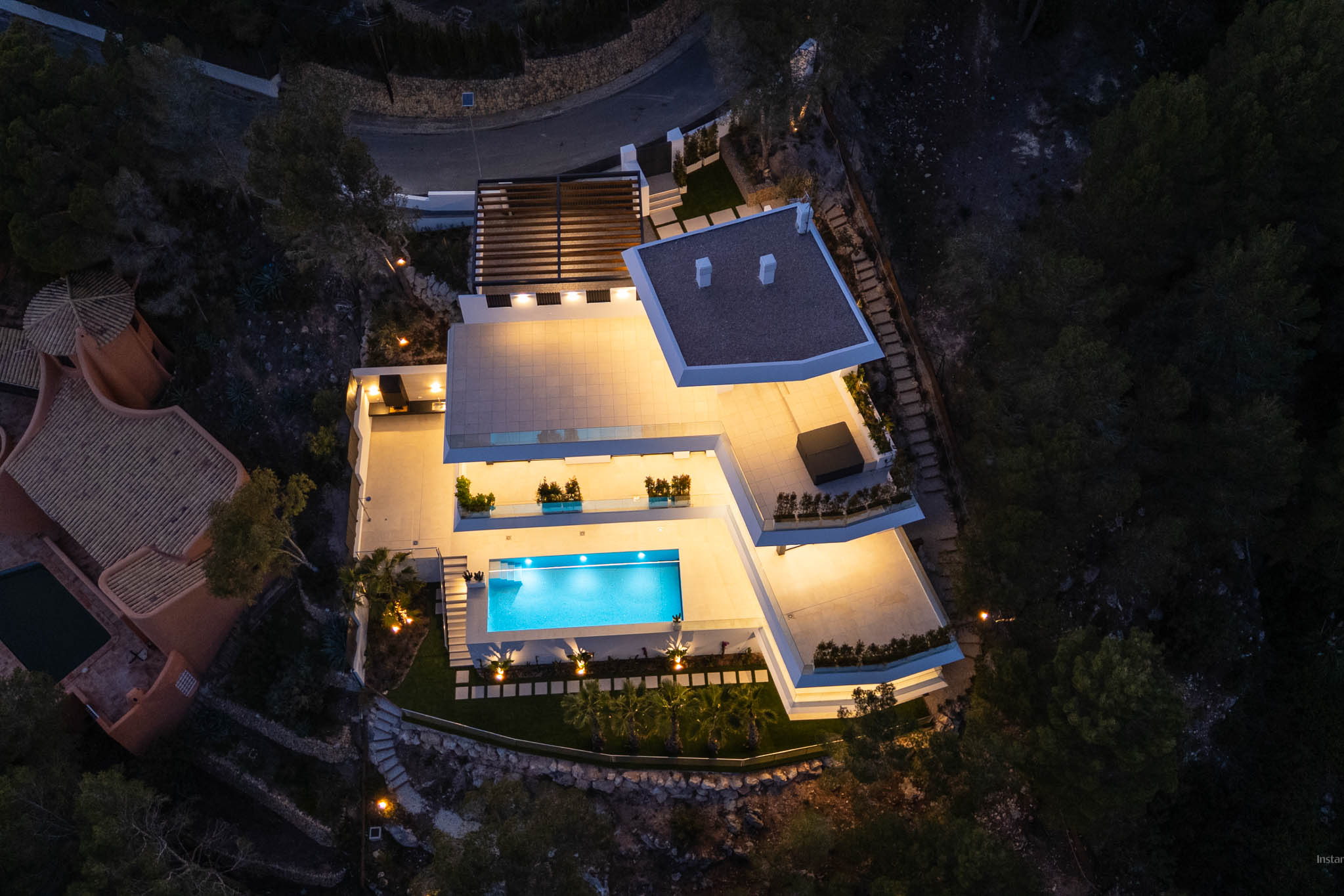 Nachtaufnahme der modernen Villa aus der Vogelperspektive, mit hell beleuchteten Terrassen und dem einladenden, blau leuchtenden Pool. Mehrere Außenwohnbereiche sind sichtbar, eingebettet in die dunkle Naturlandschaft.