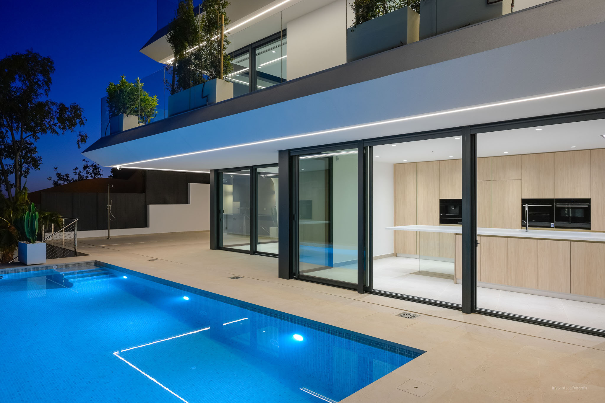 Nachtaufnahme der modernen Villa mit beleuchtetem blauen Pool und klarer Architektur. Eine helle Holzküche ist durch die großen Glasfronten sichtbar.