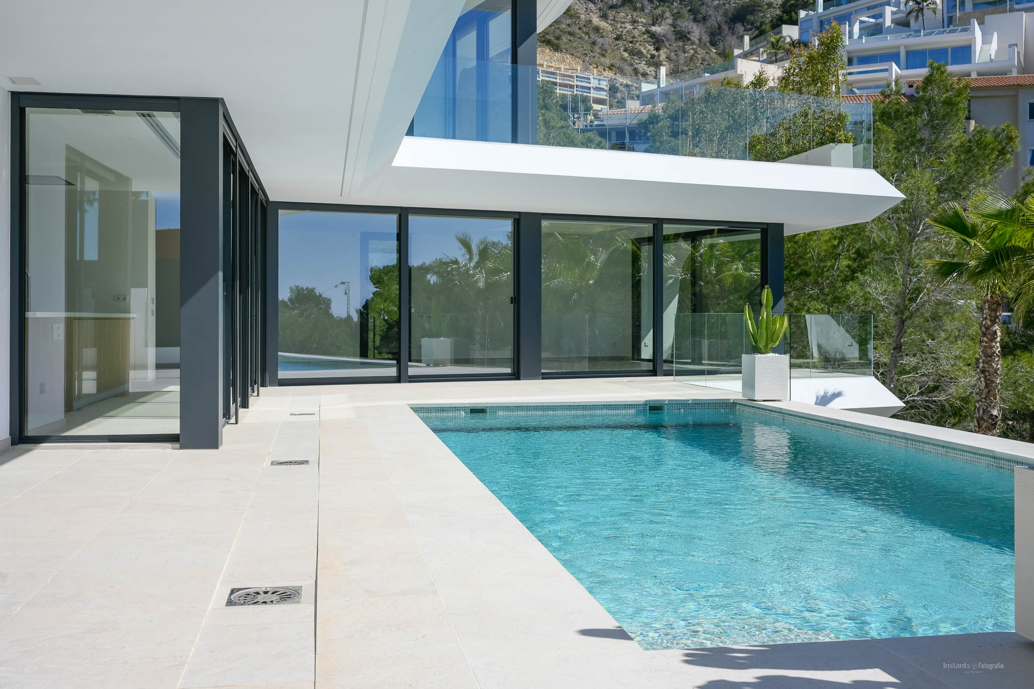 Privater Pool mit blauen Mosaikfliesen und heller Steinterrasse, umrahmt von großen Glastüren, die zum modernen Innenbereich der Villa führen.
