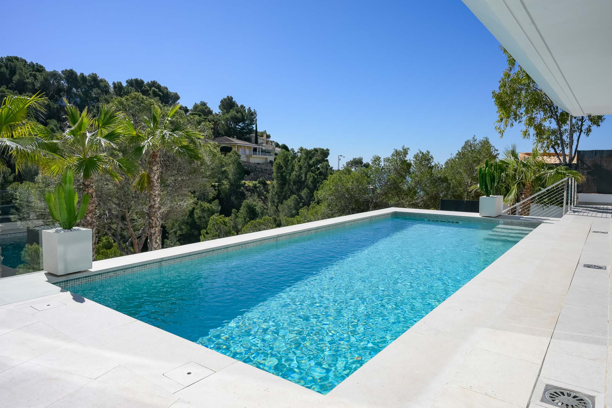 Rechteckiger Pool mit hellblauen Mosaikfliesen und weißen Steinumrandungen, umgeben von mediterraner Vegetation unter klarem Himmel.