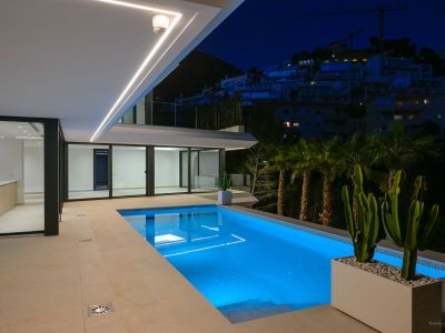 Beleuchteter Pool bei Nacht mit blauem Unterwasserlicht und gefliester Terrasse. Moderne lineare Deckenleuchten betonen die Architektur, mit Blick auf einen Hügel und entfernte Gebäude.
