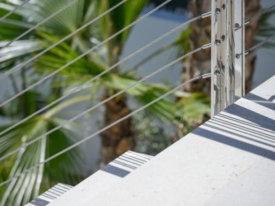 Detail einer modernen Außentreppe: helle Stufen, poliertes Metallgeländer mit Stahlseilen. Sonnenlicht wirft scharfe parallele Schatten, umrahmt von üppigem Palmenlaub.