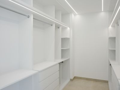Geräumiger begehbarer Kleiderschrank mit maßgefertigten weißen Schränken, verchromten Kleiderstangen und integrierter LED-Beleuchtung. Bietet mehrere Regale und Schubladen auf hellem Fliesenboden.