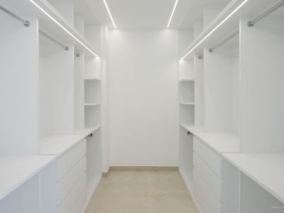 Geräumiger weißer Ankleideraum mit maßgefertigten Regalen, Schubladen und Kleiderstangen, beleuchtet durch integrierte LED-Streifen und helle Fliesen.