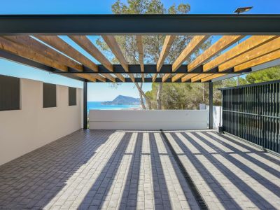 Großzügiger Außenbereich mit Pergola aus Holzlatten und Metallrahmen auf Kopfsteinpflaster. Bietet einen Panoramablick auf Meer und Berge, mit markanten Schatten durch das natürliche Licht.