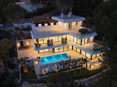 Luftaufnahme der modernen, mehrstöckigen Villa in der Dämmerung, mit beleuchteten Terrassen, einem hellblauen Pool und einer Außenküche, elegant beleuchtet inmitten üppiger Vegetation.
