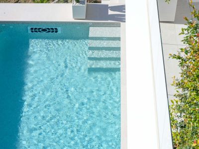 Luftaufnahme des hellblauen Mosaikpools mit integrierten Stufen, einem modernen Wasserrücklaufsystem und umliegendem hellem Pflaster.