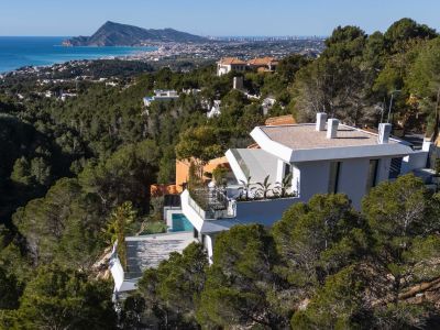Luftaufnahme einer modernen weißen Villa an einem pinienbewachsenen Hang, mit Panoramablick auf das Mittelmeer und die Küste von Altea. Ein privater Pool ist auf einer unteren Terrasse sichtbar.
