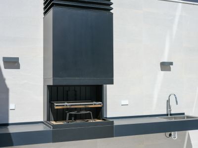 Moderne Außenküche mit dunklem Einbaugrill und Abzugshaube, passender Arbeitsplatte mit Edelstahlspüle und Chromarmatur, vor einer hell gefliesten Wand.