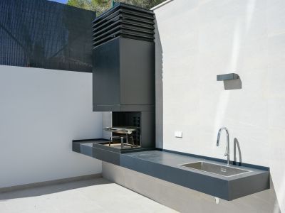 Moderne Außenküche mit integriertem dunkelgrauem Grill, passender Arbeitsplatte und Edelstahlspüle. Eine Chromarmatur ergänzt die helle Fliesenwand und den Boden.