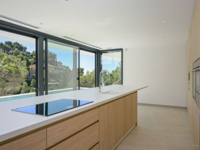 Moderne Küche mit großer Insel, weißer Arbeitsplatte, integriertem Induktionskochfeld und Chromarmatur. Schiebetüren bieten Blick auf mediterrane Vegetation und einen privaten Pool.