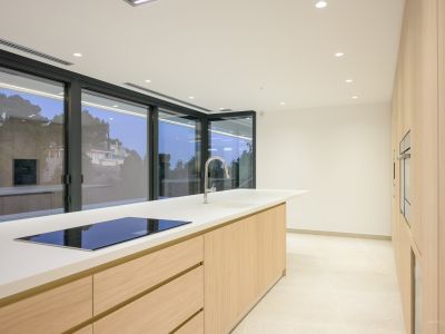 Moderne Küche mit großer Mittelinsel, weißer Arbeitsplatte und integriertem Induktionskochfeld. Helle Holzschränke und Einbauöfen an der Wand, ergänzt durch raumhohe Fenster.
