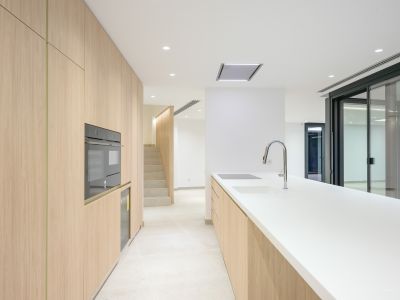 Moderne Küche mit hellen Holzschränken, weißer Arbeitsplatte und integrierten Geräten wie Backofen, Induktionskochfeld und Spüle mit Chromarmatur. Verbindung zu Treppe und Außenzugang.