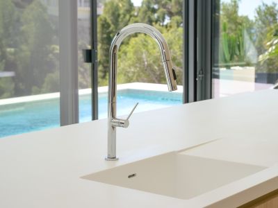 Moderne Küche mit heller Arbeitsplatte und integriertem Spülbecken. Ein eleganter Chrom-Ausziehhahn blickt auf den Pool und die grüne Außenanlage durch große Glasfronten.