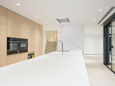 Moderne Küche mit langer weißer Insel, integrierter Spüle und Chromarmatur, ergänzt durch helle Holzschränke bis zur Decke und integrierte Doppelöfen. Einbauleuchten erhellen den zeitgenössischen Raum.
