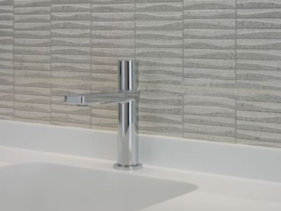 Moderner Chromwasserhahn auf einer weißen Arbeitsplatte mit integriertem Waschbecken. Die Wand ist mit strukturierten hellen Fliesen im Wellenmuster versehen.