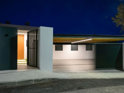 Moderner Eingangsbereich mit dunklem Fußgängertor und Holztür, daneben ein beleuchteter Carport mit Lamellendecke und linearer LED-Beleuchtung.