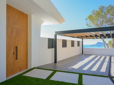 Moderner Eingangsbereich mit hoher Holztür, Kunstrasenweg und gepflastertem Parkplatz unter einer Metallpergola. Im Hintergrund sind das Mittelmeer und Berge sichtbar.