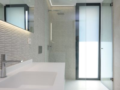Modernes Badezimmer mit weißem Waschbecken, Chromarmatur und strukturierten grauen Wandfliesen. Es verfügt über eine Glasdusche mit Regenduschkopf und LED-beleuchteten Spiegel.