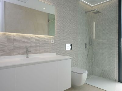 Modernes Badezimmer mit weißem Waschtisch, beleuchtetem Spiegel und strukturierten grauen Wandfliesen. Wandhängende Toilette und geräumige Duschkabine mit Glaswand.