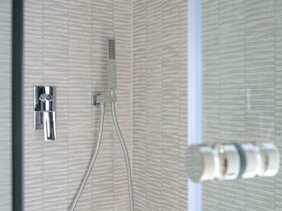 Modernes Duschdetail mit grauen, strukturierten Rechteckfliesen, einer eleganten Chrom-Mischbatterie und Handbrause. Eine Glasduschwand ist teilweise sichtbar.