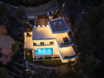 Nachtaufnahme der modernen Villa aus der Vogelperspektive, mit hell beleuchteten Terrassen und dem einladenden, blau leuchtenden Pool. Mehrere Außenwohnbereiche sind sichtbar, eingebettet in die dunkle Naturlandschaft.