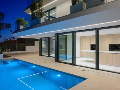 Nachtaufnahme der modernen Villa mit beleuchtetem blauen Pool und klarer Architektur. Eine helle Holzküche ist durch die großen Glasfronten sichtbar.