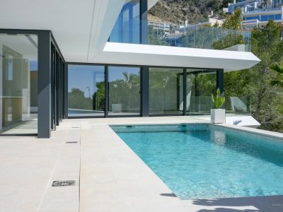 Privater Pool mit blauen Mosaikfliesen und heller Steinterrasse, umrahmt von großen Glastüren, die zum modernen Innenbereich der Villa führen.