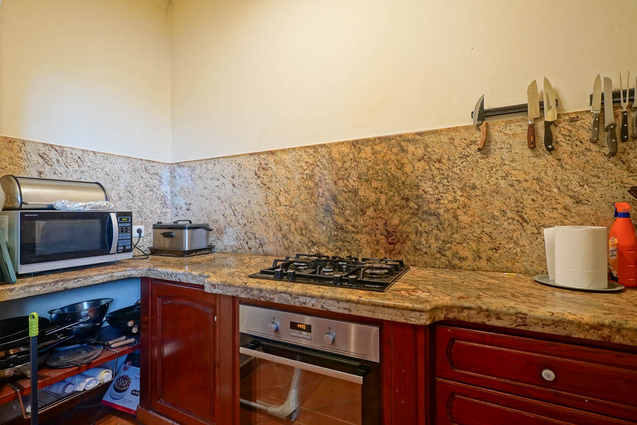Ausgestattete Küchenecke mit Arbeitsplatten und Spritzschutz aus geädertem Granit, Einbaubackofen, Gasherd und dunklen Holzschränken. Offene Regale sind sichtbar.