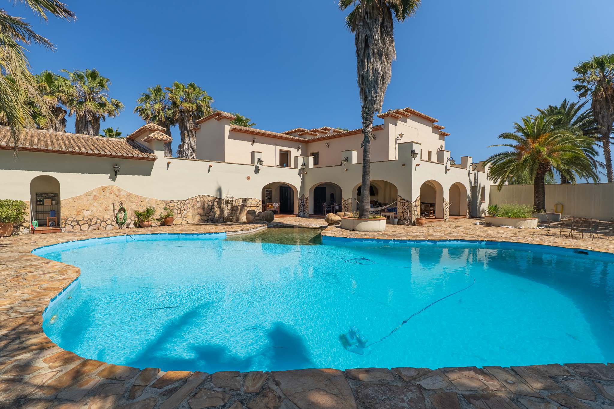 Außenansicht der mediterranen Villa mit unregelmäßig geformtem Privatpool, Natursteinumrandung und zahlreichen Palmen unter klarem blauem Himmel.