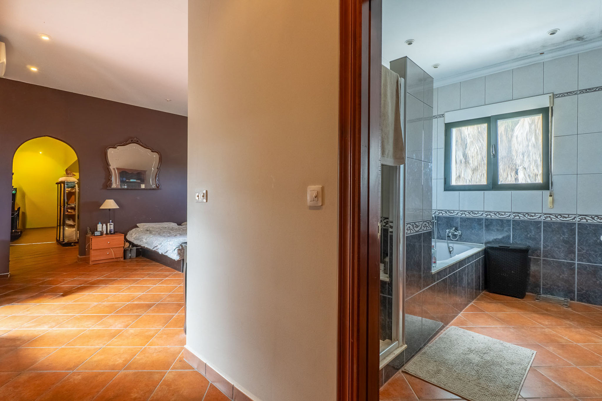 Blick vom Flur mit Terrakottafliesen, links ein Schlafzimmer mit dunklen Wänden und Zierspiegel, rechts ein gefliestes Badezimmer mit Badewanne.