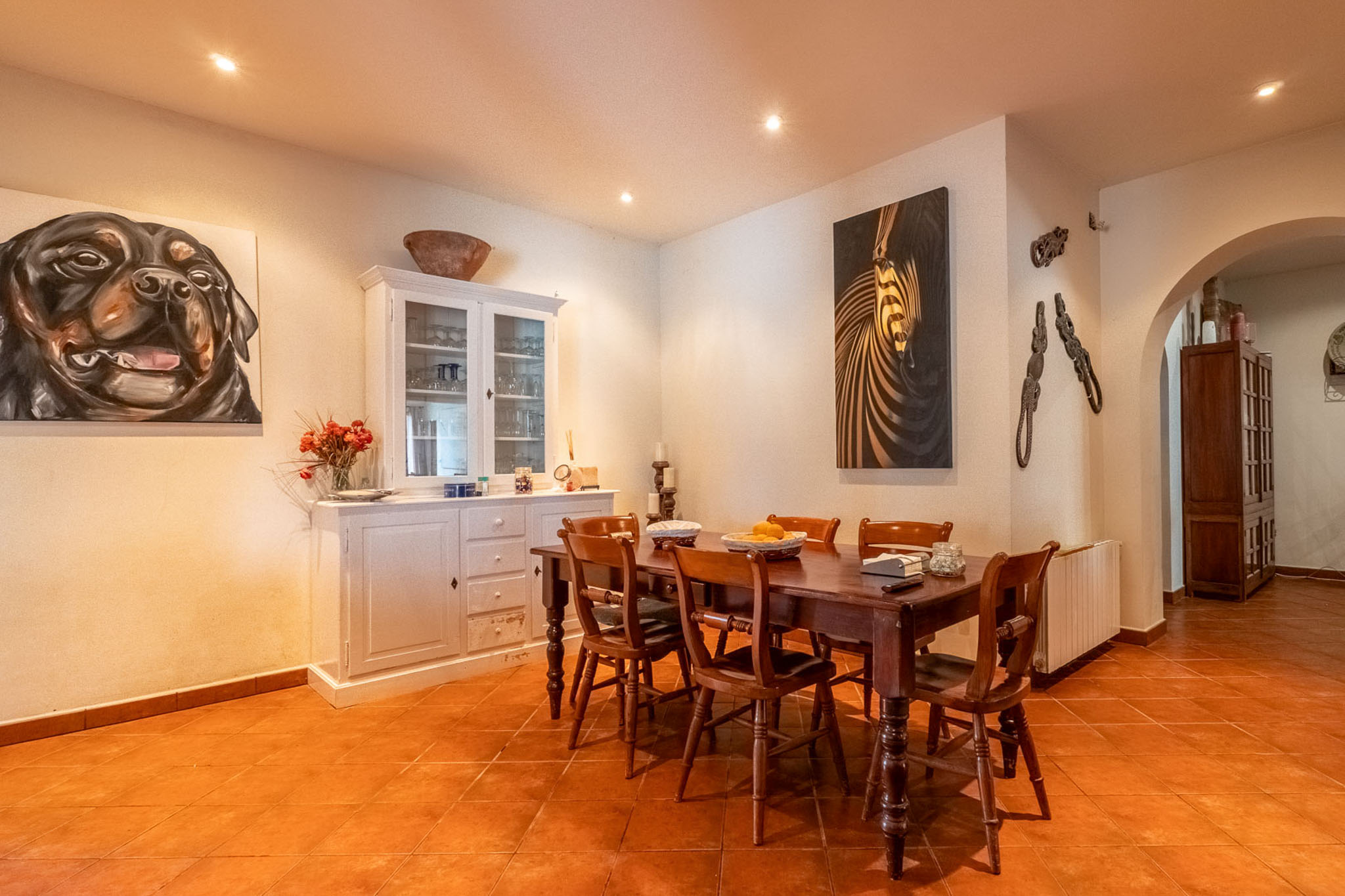 Esszimmer mit dunklem Holztisch und sechs Stühlen, Terrakotta-Fliesenboden und weißen Wänden mit Einbauleuchten. Ein weißer Schrank und zwei große Gemälde schmücken den Raum.