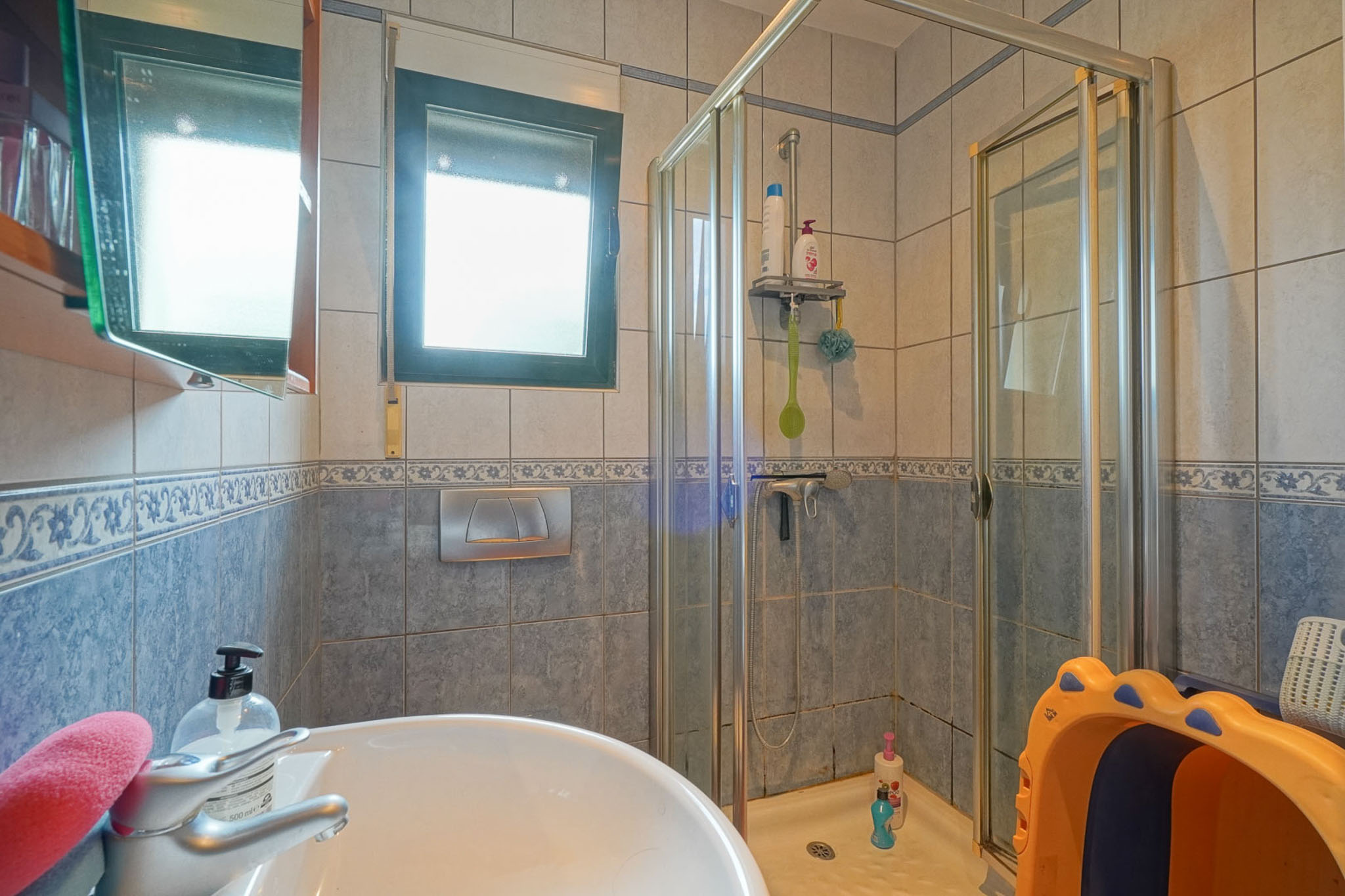 Funktionales Badezimmer mit Keramikwaschbecken, Glasduschkabine und blau-beigen Fliesen. Ein Fenster sorgt für natürliches Licht.