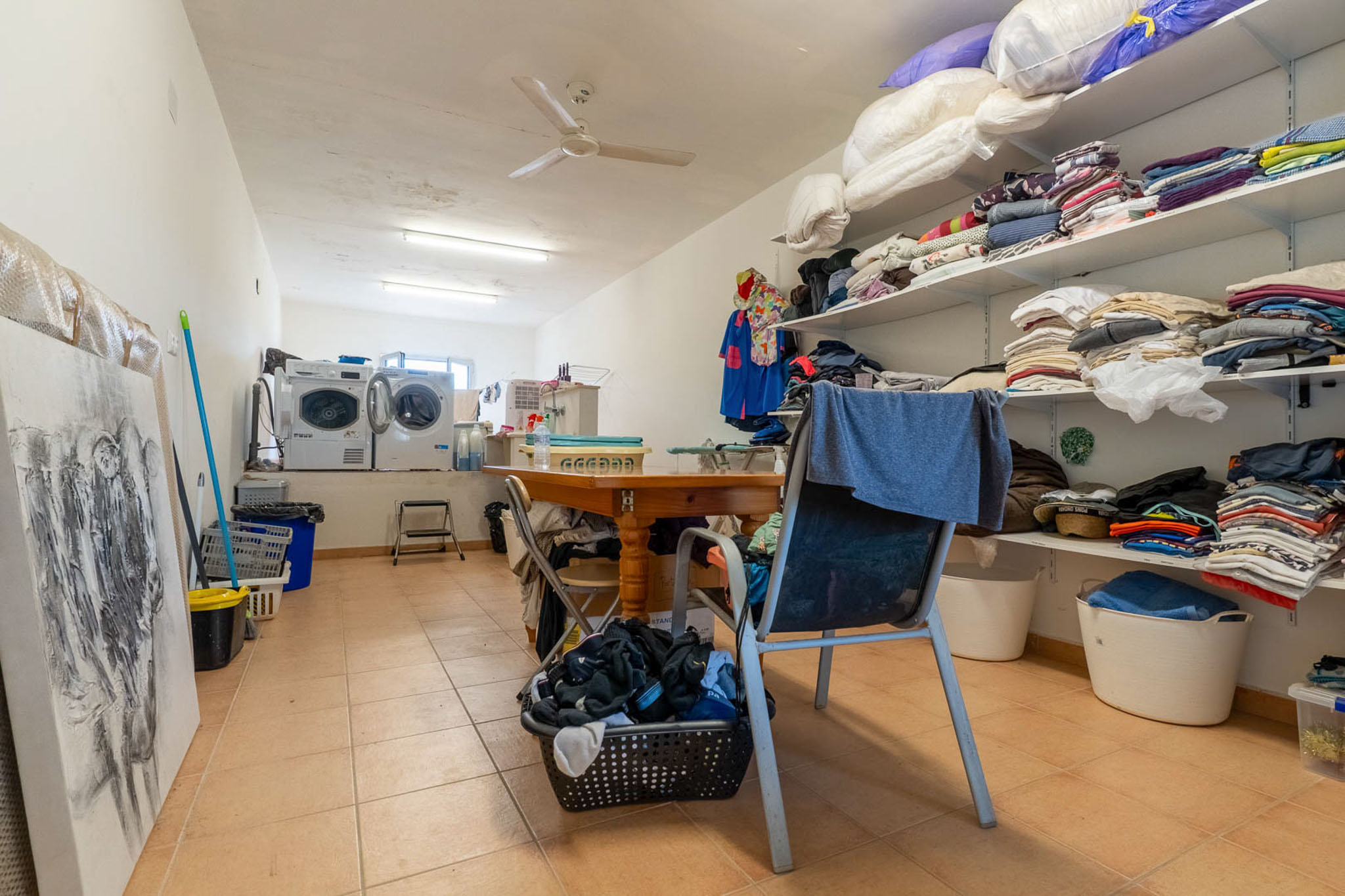 Funktioneller Hauswirtschaftsraum mit zwei Waschmaschinen, Trockner und offenen Regalen für Wäsche. Ein zentraler Holztisch bietet eine praktische Arbeitsfläche, ergänzt durch Fliesenboden und Deckenventilator.