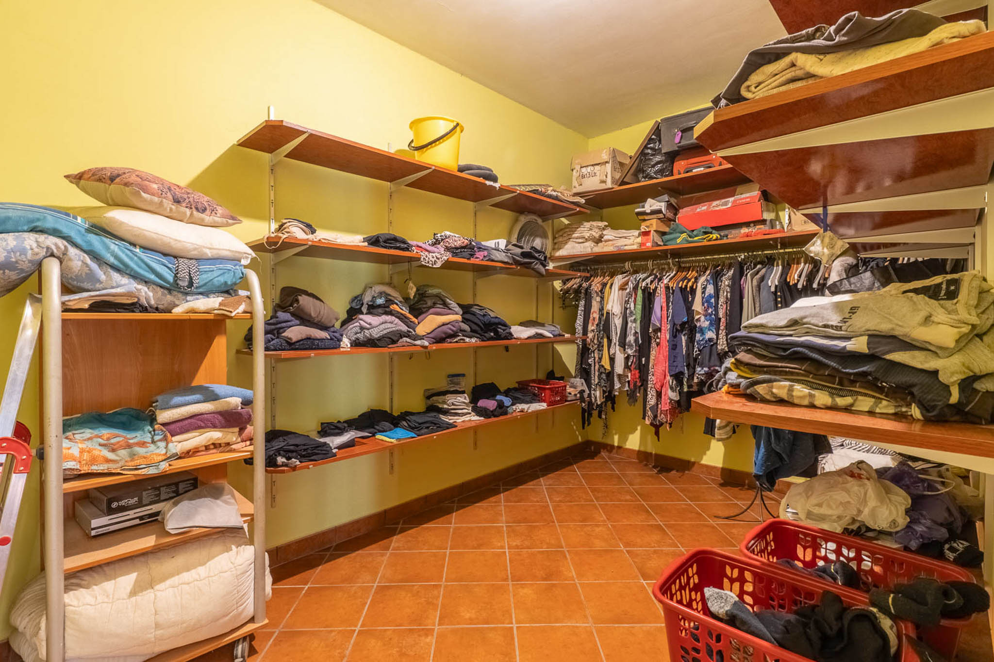 Geräumiger begehbarer Kleiderschrank mit offenen Holzregalen, hellgelben Wänden und Terrakottafliesenboden, ideal für umfassende Aufbewahrung.