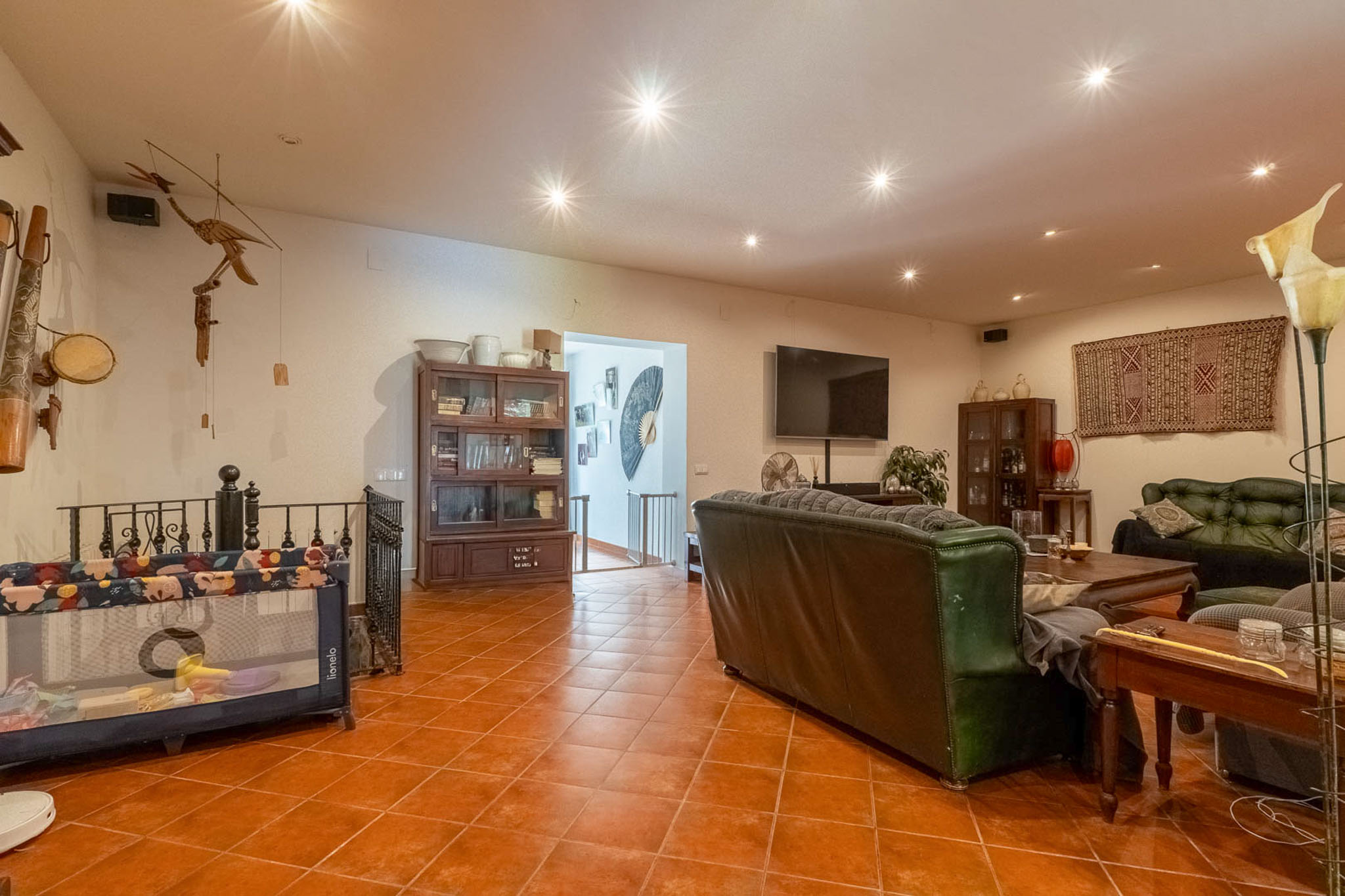 Geräumiger Wohnbereich mit Terrakotta-Fliesenboden, Einbauleuchten, dunklen Ledersofas und Holzvitrinen. Ein Wandfernseher und ein offener Zugang zum Treppenhaus sind sichtbar.