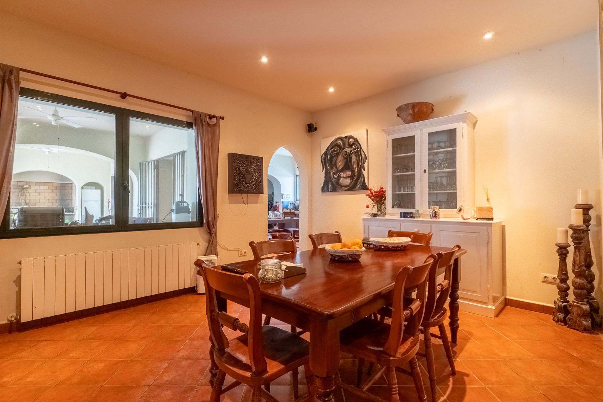 Geräumiges Esszimmer mit dunklem Holztisch und passenden Stühlen auf Terrakottafliesen. Der Raum verfügt über einen Heizkörper unter dem Fenster und einen weißen Vitrinenschrank.