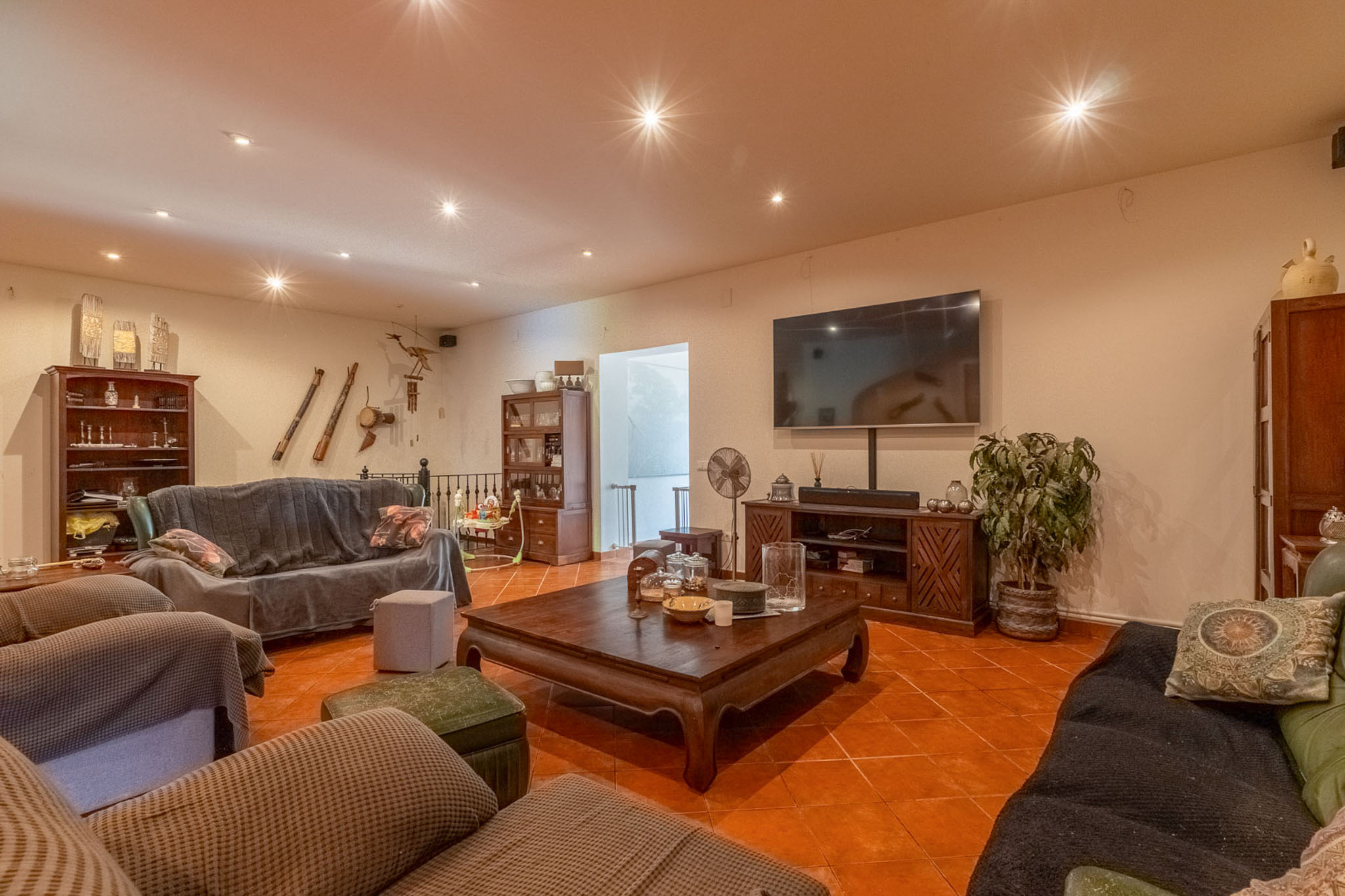 Geräumiges Wohnzimmer mit Terrakottafliesen, Deckenstrahlern und großem Wandfernseher. Dunkle Holzmöbel und überzogene Sofas schaffen ein einladendes Ambiente.
