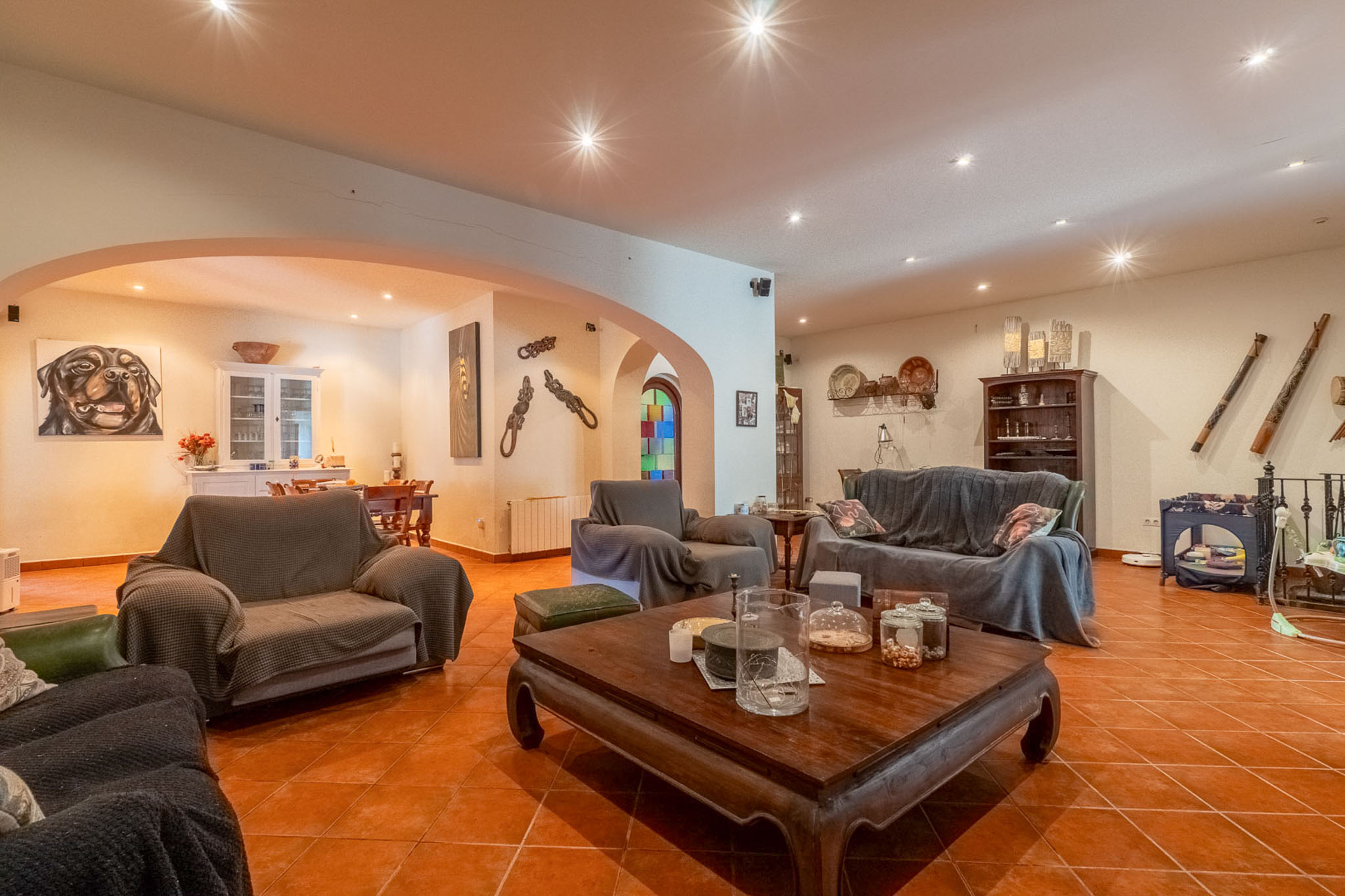 Geräumiges Wohnzimmer mit Terrakottafliesen und Deckeneinbaustrahlern. Es umfasst ein großes Sofa, Sessel und einen geschnitzten Holztisch, verbunden mit einem Essbereich durch Bögen.