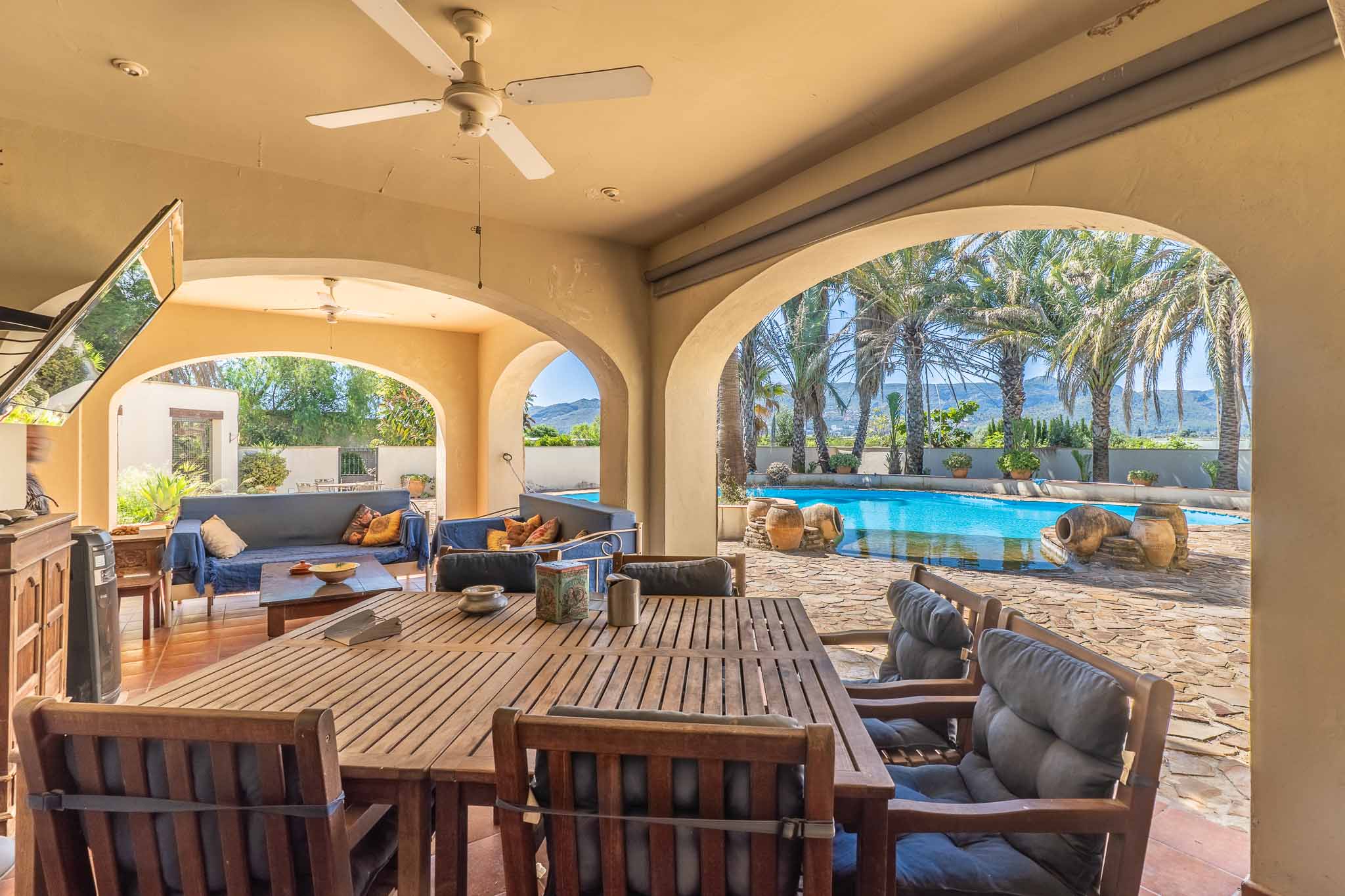 Großzügige überdachte Terrasse mit Bögen, ausgestattet mit Holz-Esstisch und Loungebereich, mit direktem Blick auf den Pool, Palmen und die Berglandschaft.