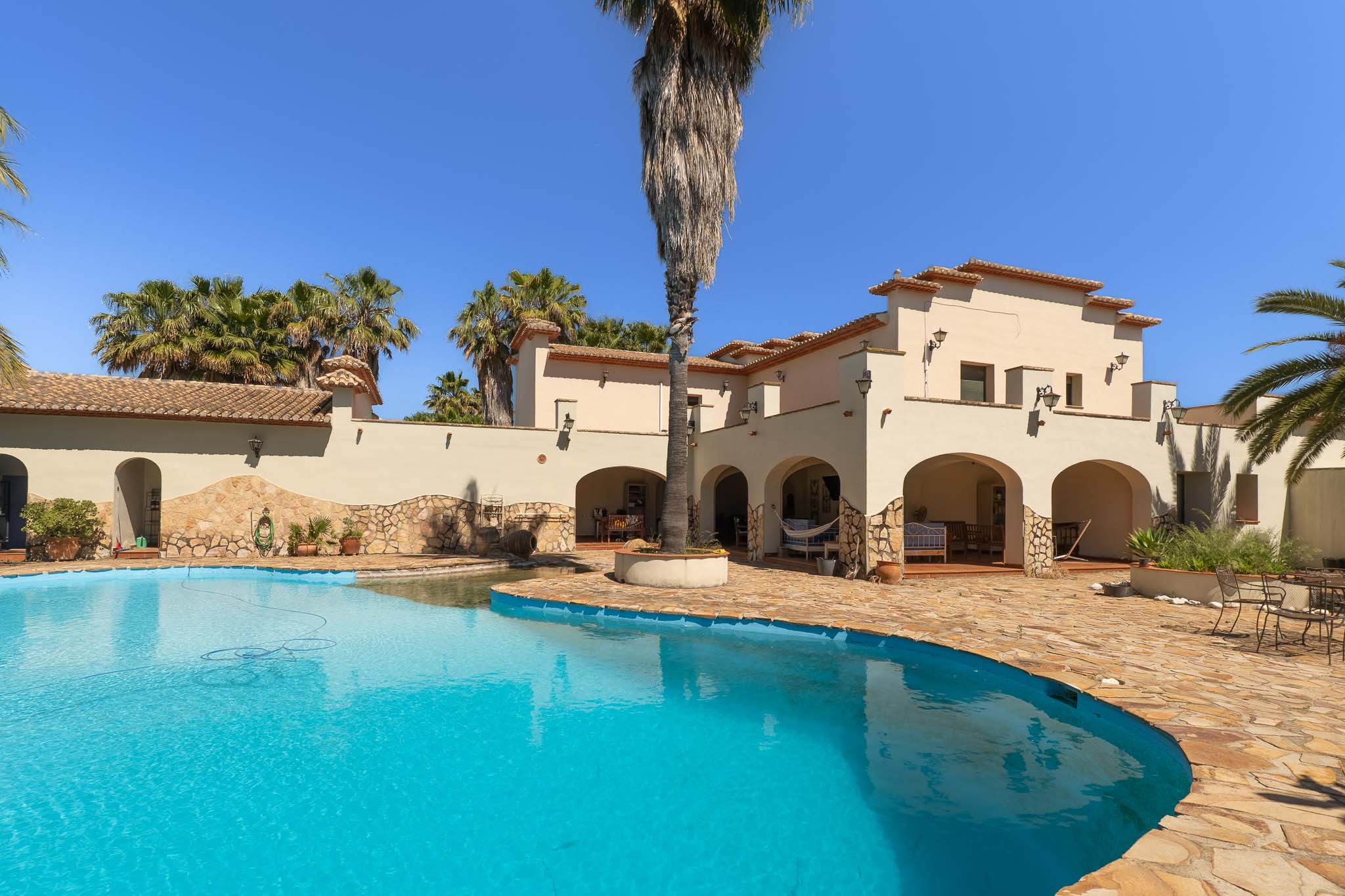 Großzügiger, unregelmäßig geformter Privatpool mit klarem blauen Wasser, umgeben von einer weitläufigen Natursteinterrasse. Die mediterrane Villa zeigt Bögen und Ziegeldächer inmitten von Palmen.