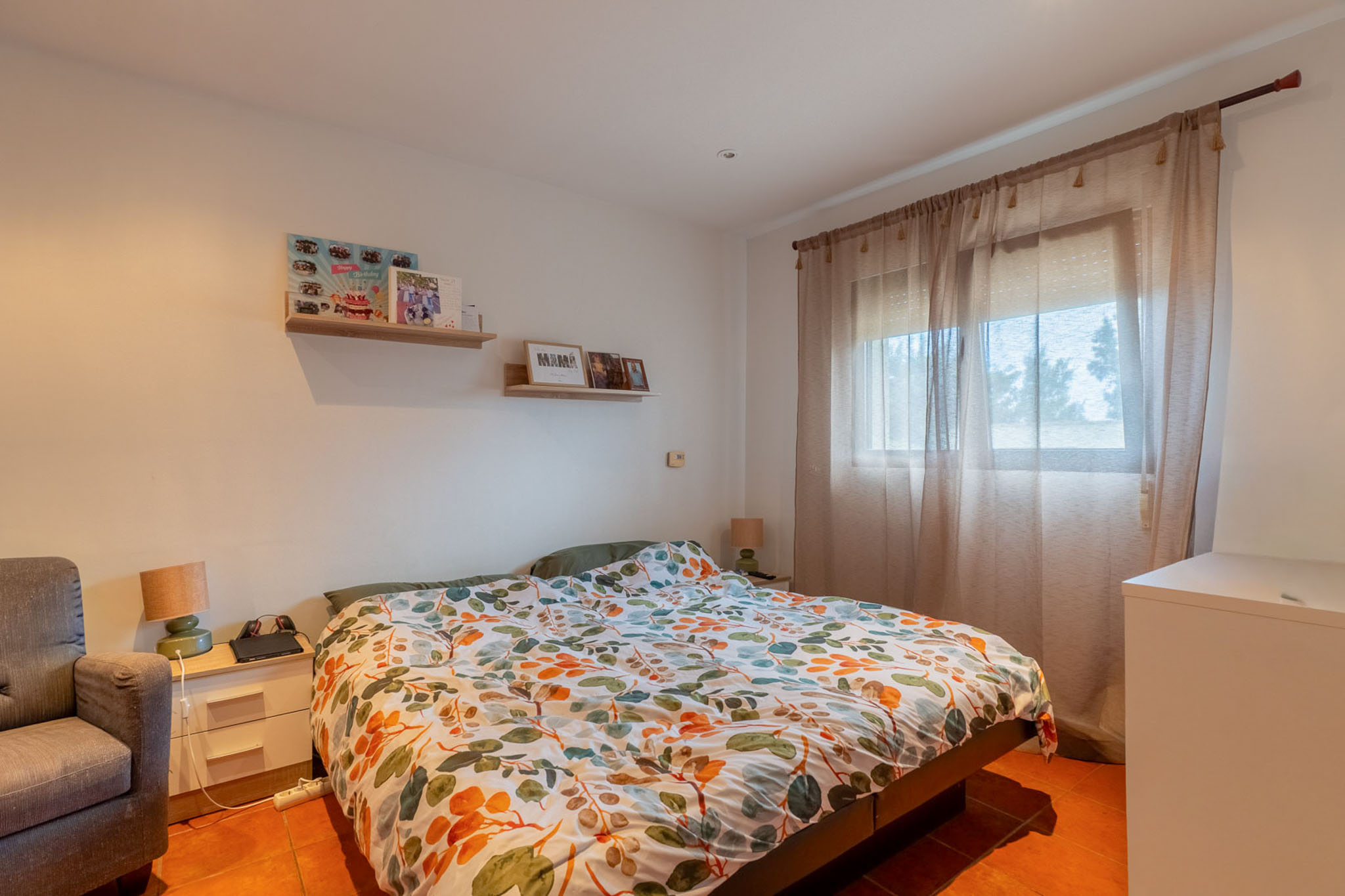 Helles Schlafzimmer mit Doppelbett, bunter Blätterbettdecke und Terrakottafliesenboden. Natürliches Licht fällt durch ein Fenster mit Gardinen, ergänzt durch Nachttisch und Wandregale.