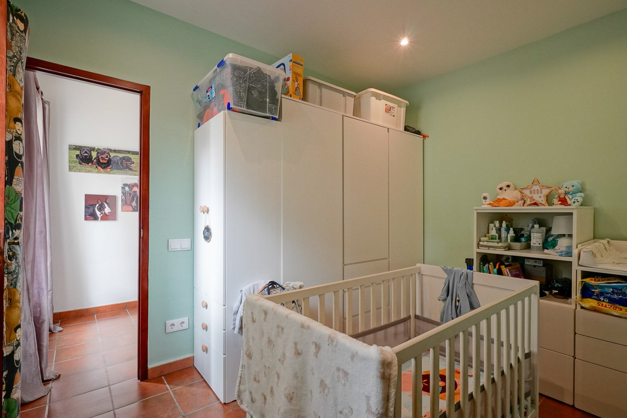 Kinderzimmer mit hellgrünen Wänden und Terrakottafliesenboden. Ausgestattet mit einem weißen Babybett, geräumigen weißen Kleiderschränken und einem Regal, beleuchtet durch Deckenstrahler.