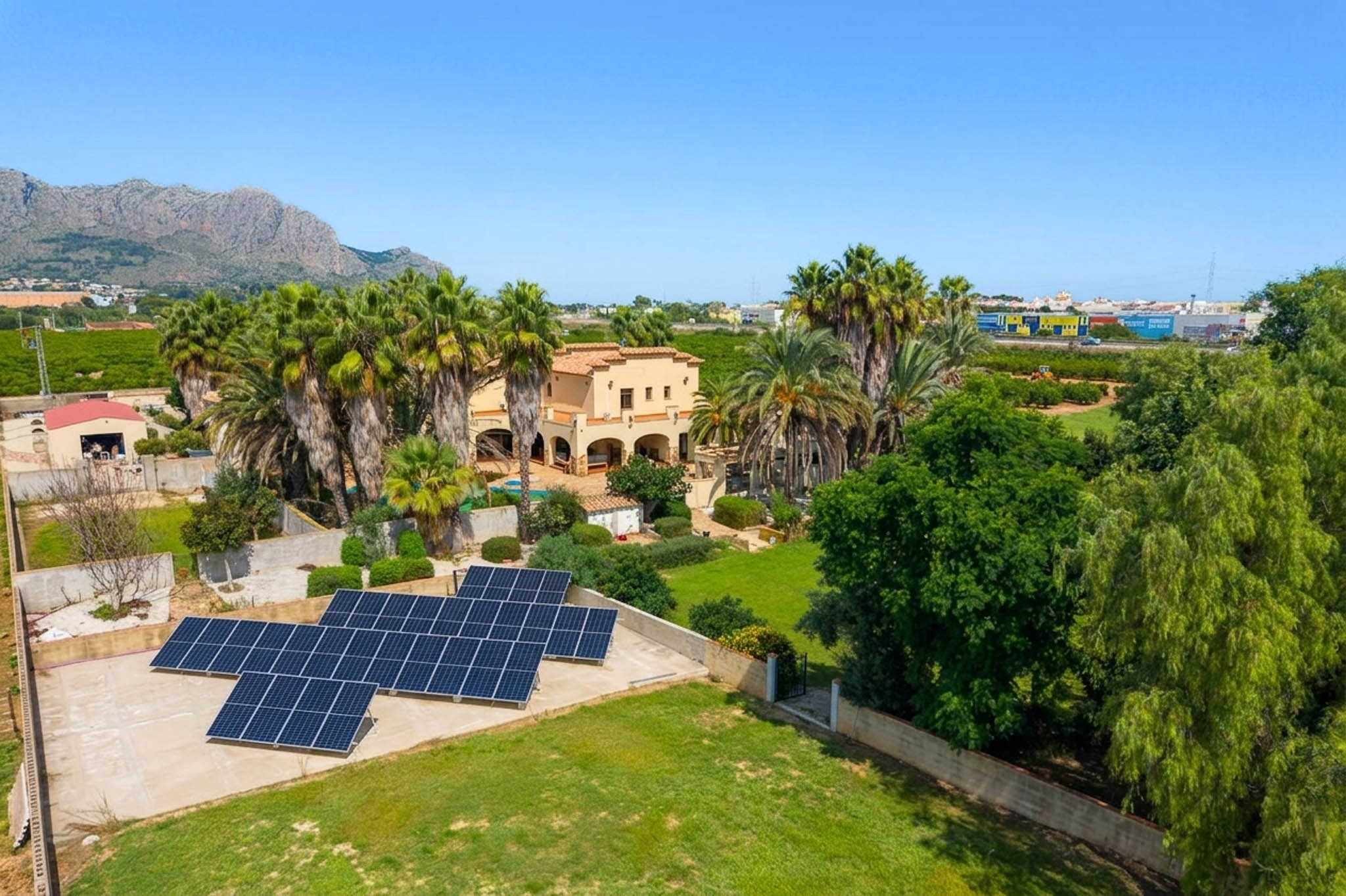 Luftaufnahme der traditionellen Villa, umgeben von Palmen und üppigen Gärten, mit einer prominenten Solaranlage und fernen Bergblicken unter klarem Himmel.