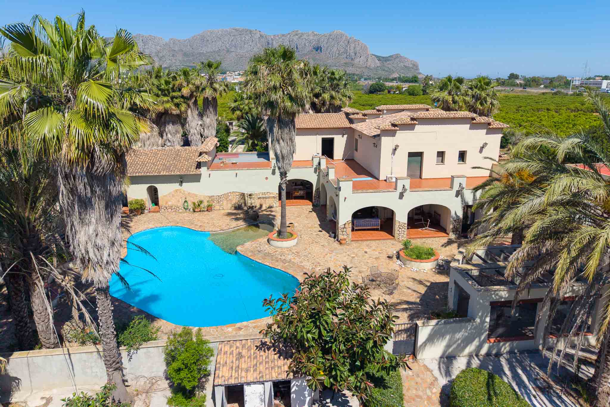 Luftaufnahme der Villa mit großem Freiformpool, weitläufigem gepflasterten Patio und Palmen, vor einer Bergkulisse und grünen Feldern.