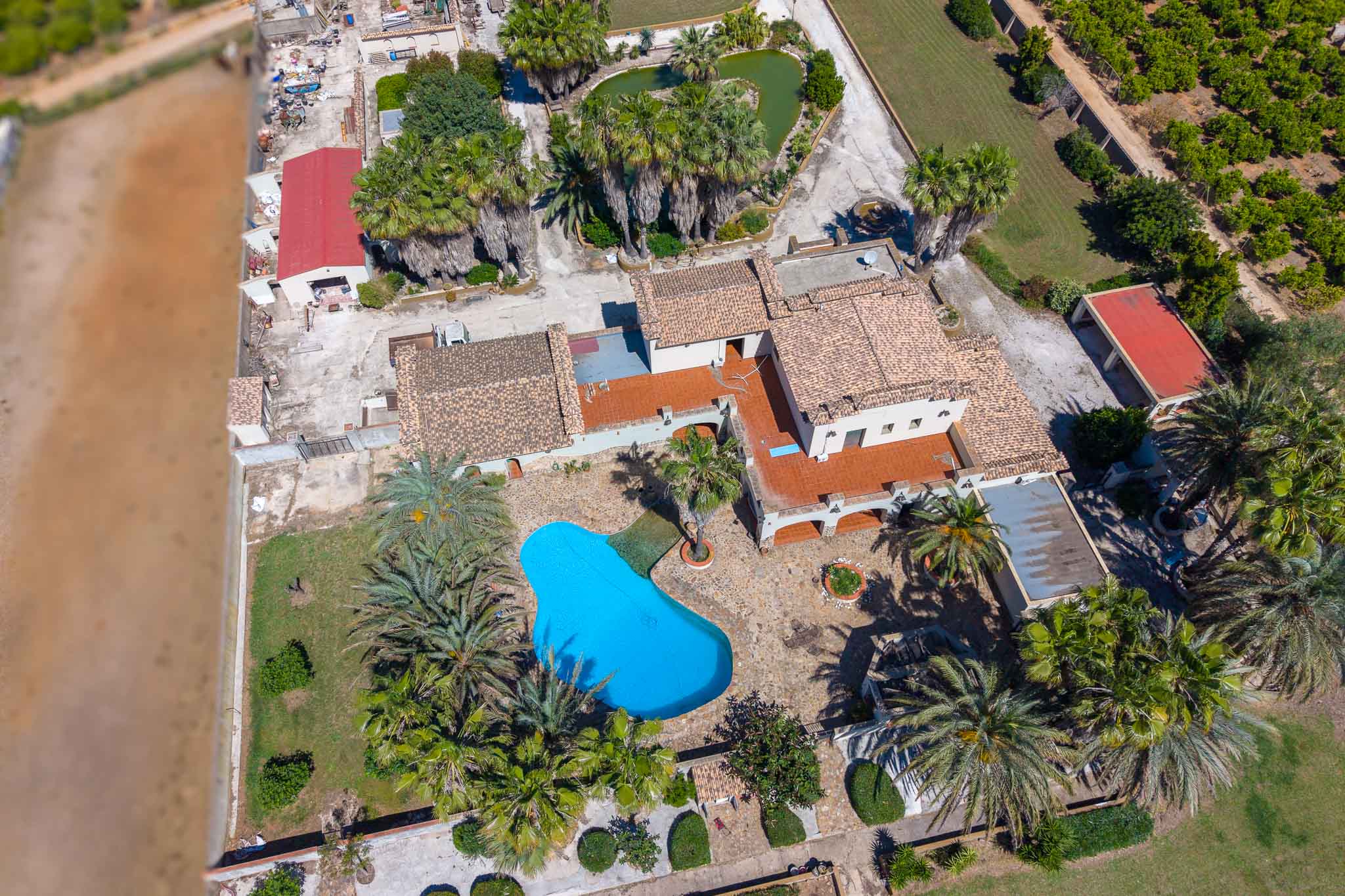 Luftaufnahme des Anwesens mit unregelmäßig geformtem blauem Pool, umgeben von Palmen und Gärten. Mehrere Gebäude mit Ziegeldächern und ein herzförmiger Teich sind sichtbar.