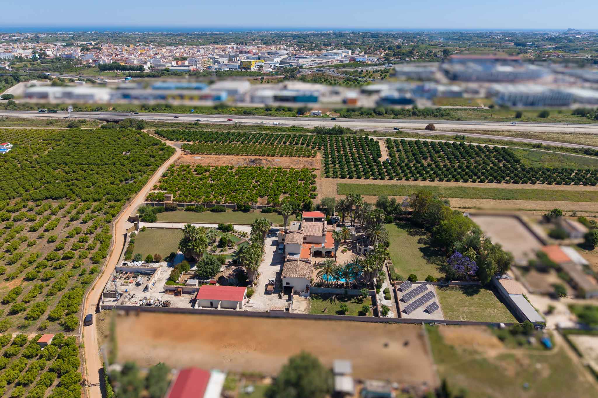 Panoramische Luftaufnahme des Anwesens, mit privatem Pool, Solarpaneelen und weitläufigen Orangenhainen. Eine Autobahn, Stadt und das ferne Mittelmeer runden die Landschaft ab.