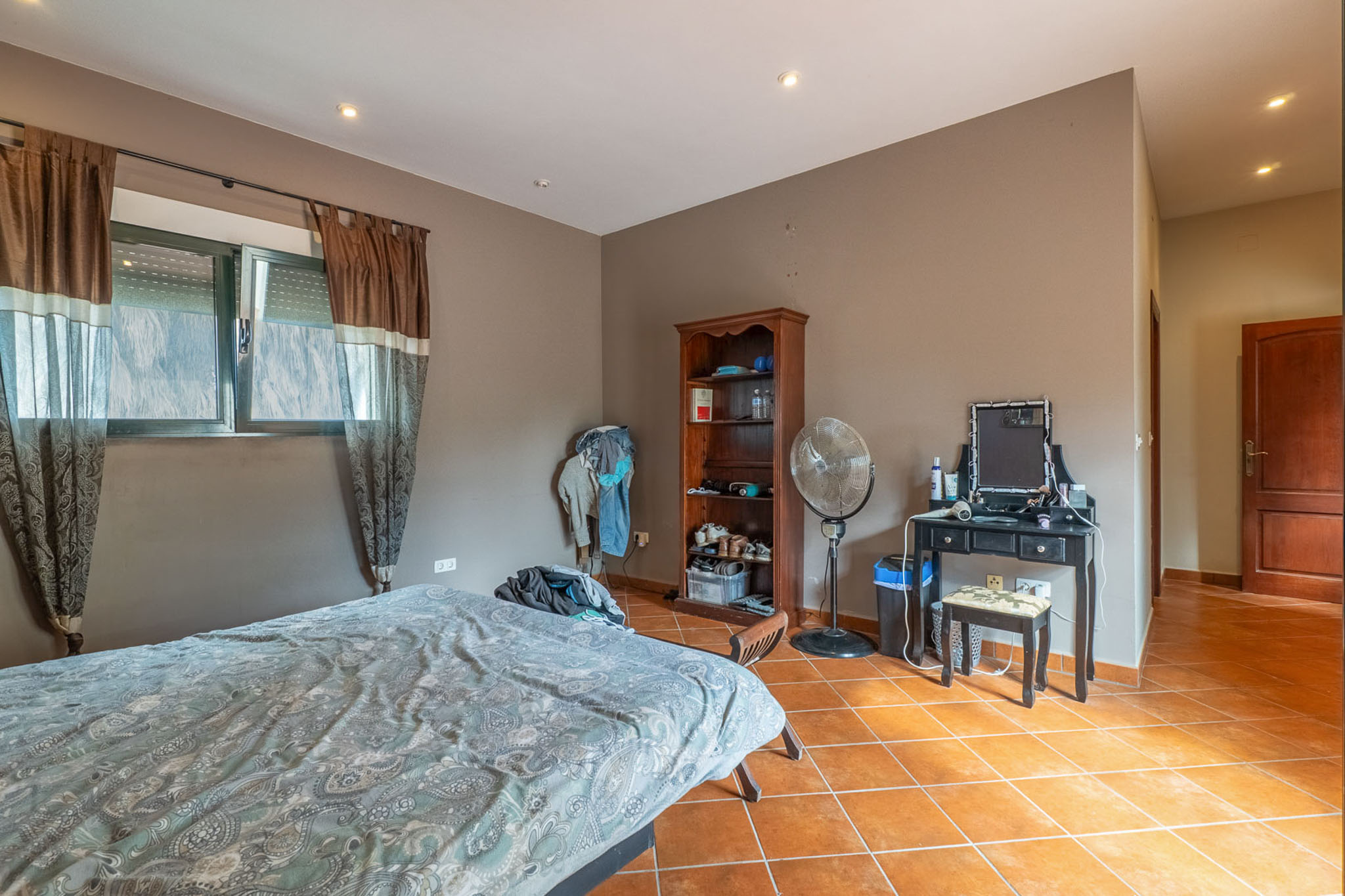 Schlafzimmer mit Bett, Fenstern mit braunen Gardinen und Rollos, sowie Terrakotta-Fliesenboden. Ausgestattet mit schwarzem Schminktisch, Holzregal und Standventilator.