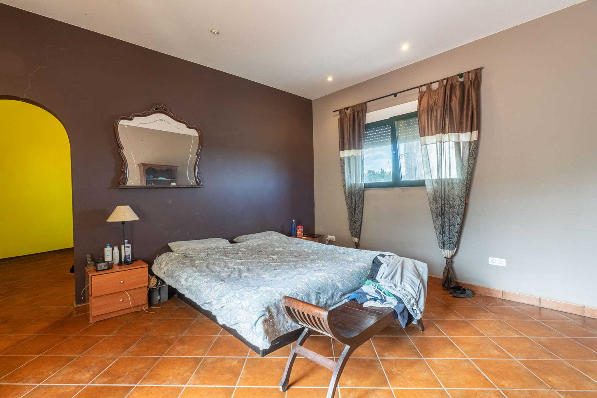 Schlafzimmer mit Doppelbett, Terrakottafliesen und zweifarbigen Wänden. Das Fenster mit Rollladen und Vorhängen sorgt für natürliches Licht.